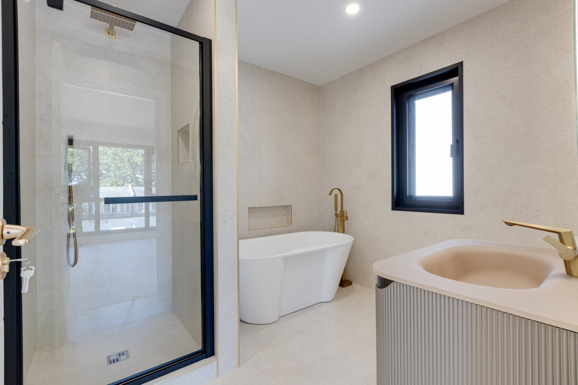 Modern beige bathroom with a glass shower, freestanding tub, ribbed vanity, black window frames, and gold fixtures.