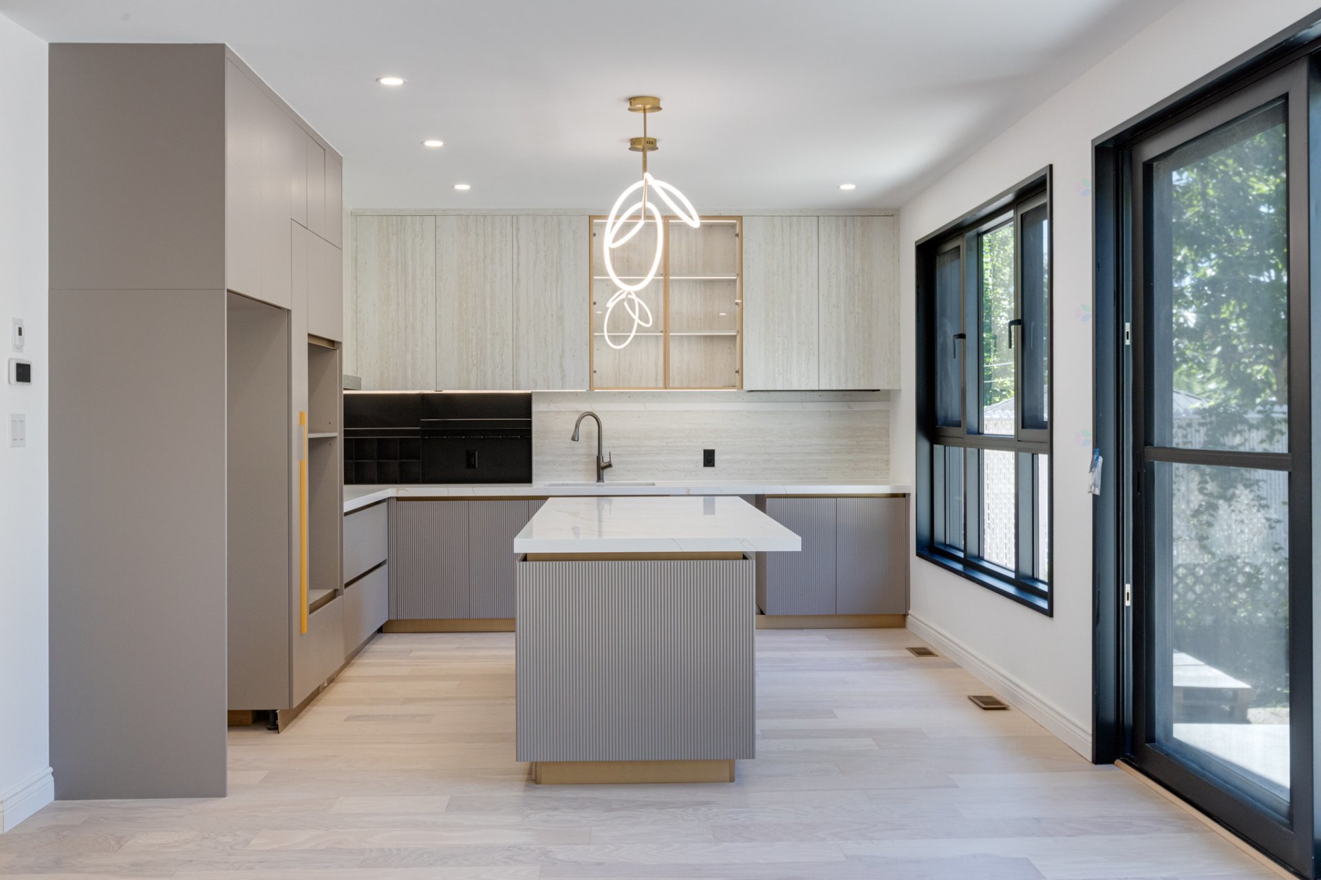 Modern light-gray kitchen with a ribbed island, sculptural pendant light, black-framed windows, and pale wood flooring.