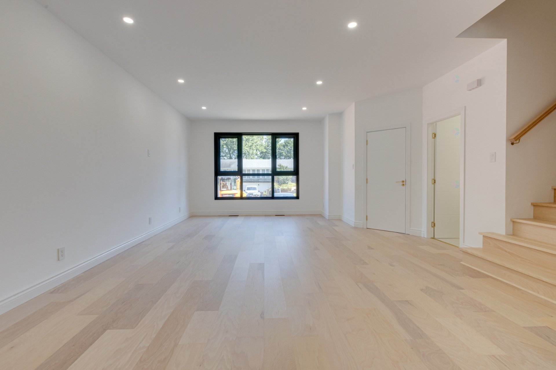Empty modern white room with pale wood floors, a large window, doors, recessed lights, and stairs.