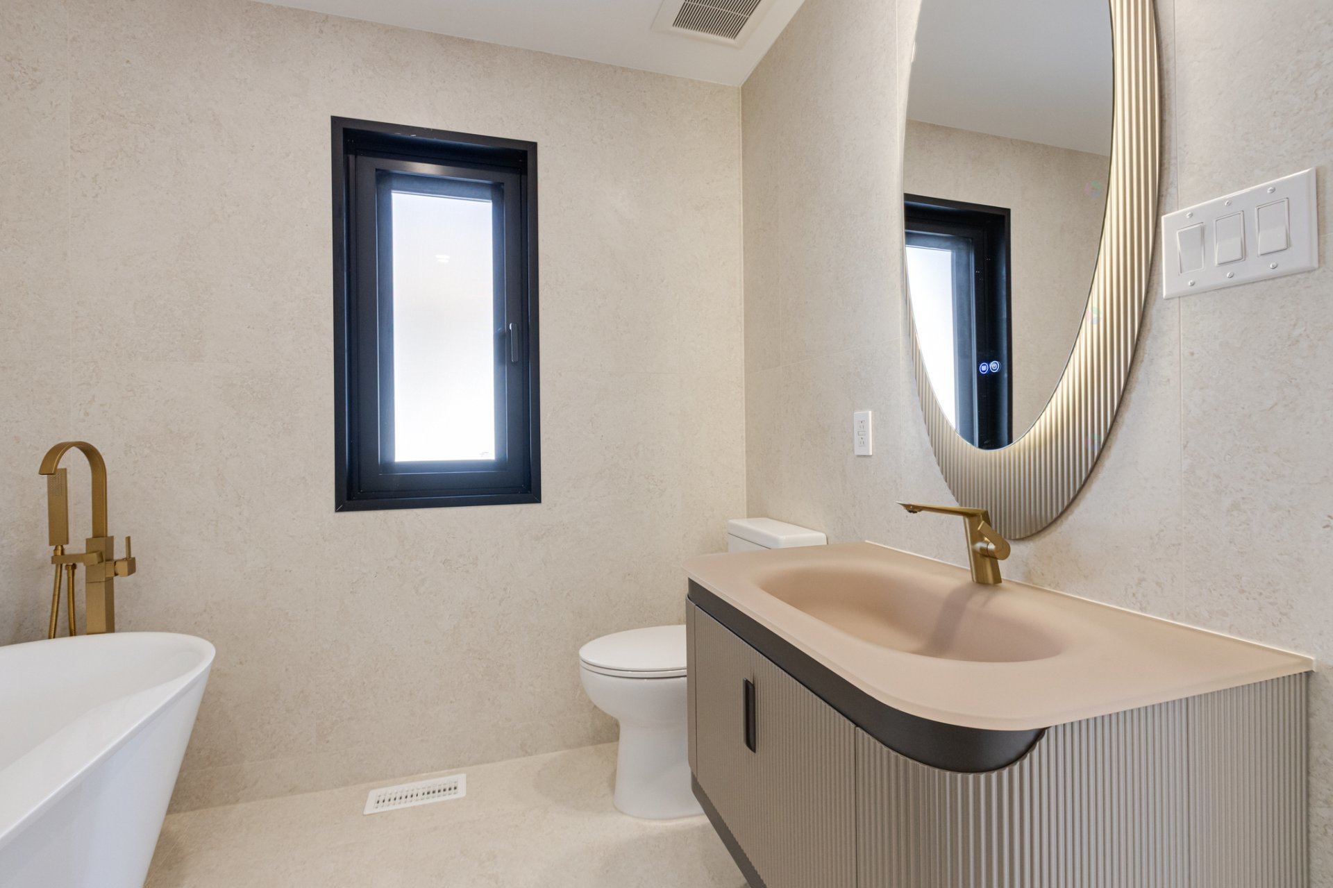 Modern beige-tiled bathroom with bathtub, toilet, ribbed vanity, gold fixtures, black-framed window, and curved mirror.
