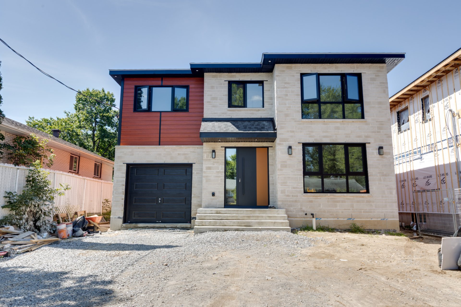 Modern two-story house with cream masonry, reddish siding, black windows, garage, and unfinished gravel frontage.